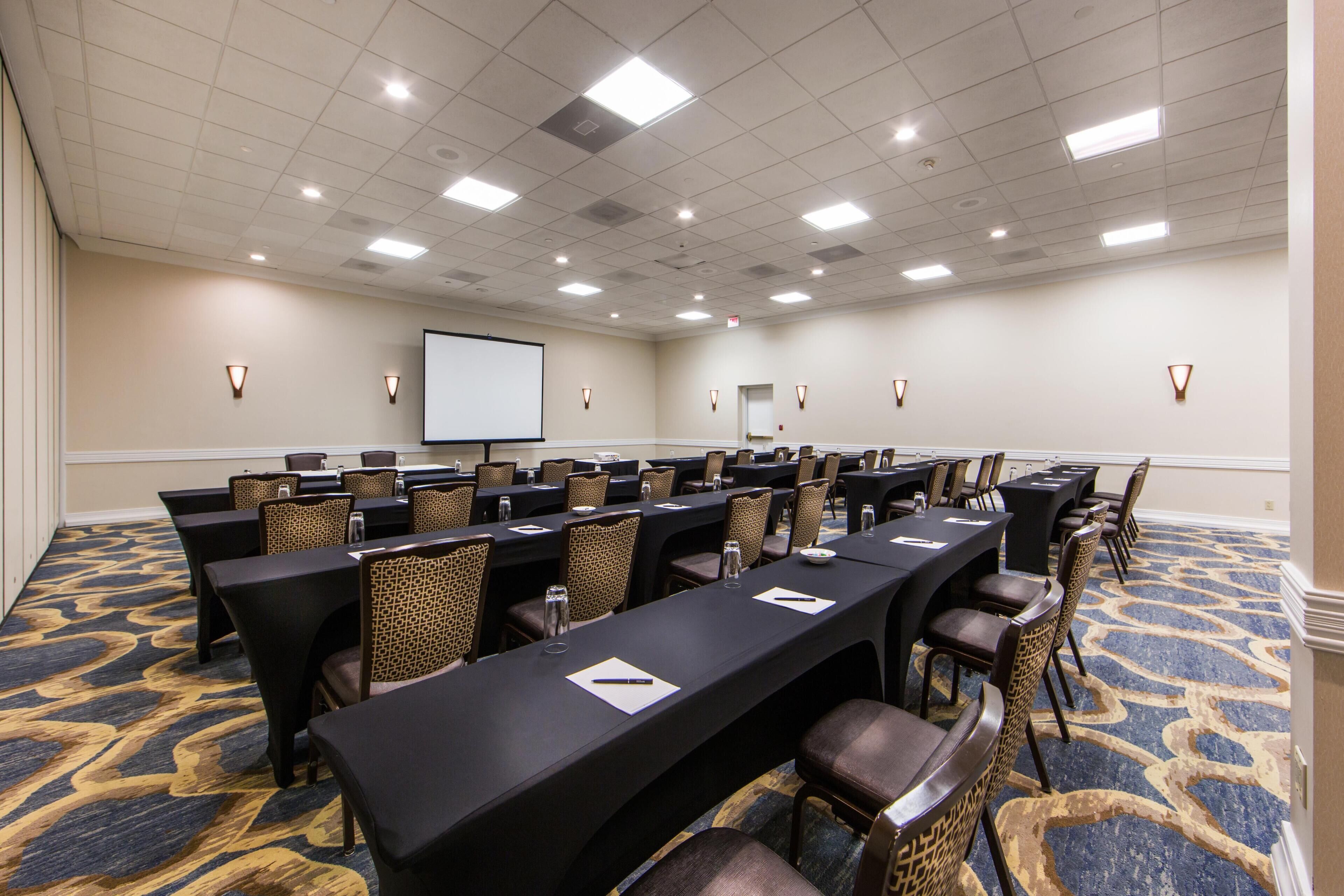 A conference room with multiple tables, chairs, a projector screen, and a carpeted floor.