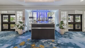 A reception area with a dark reception desk, potted plants, and a view through the glass doors. - Hilton Irvine/Orange County Airport (Irvine)