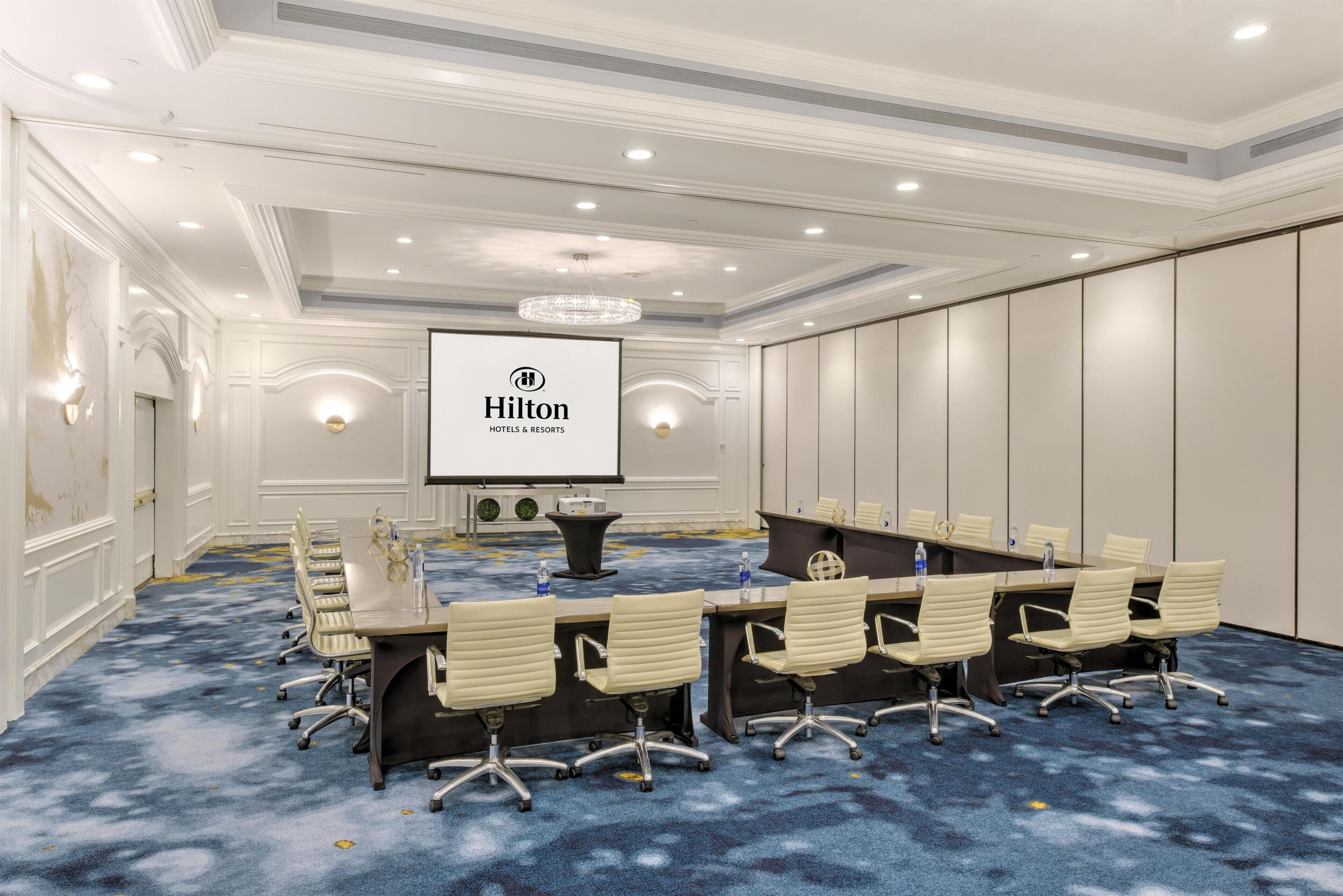 A conference room with a long table, chairs, a projector screen, and a carpeted floor.