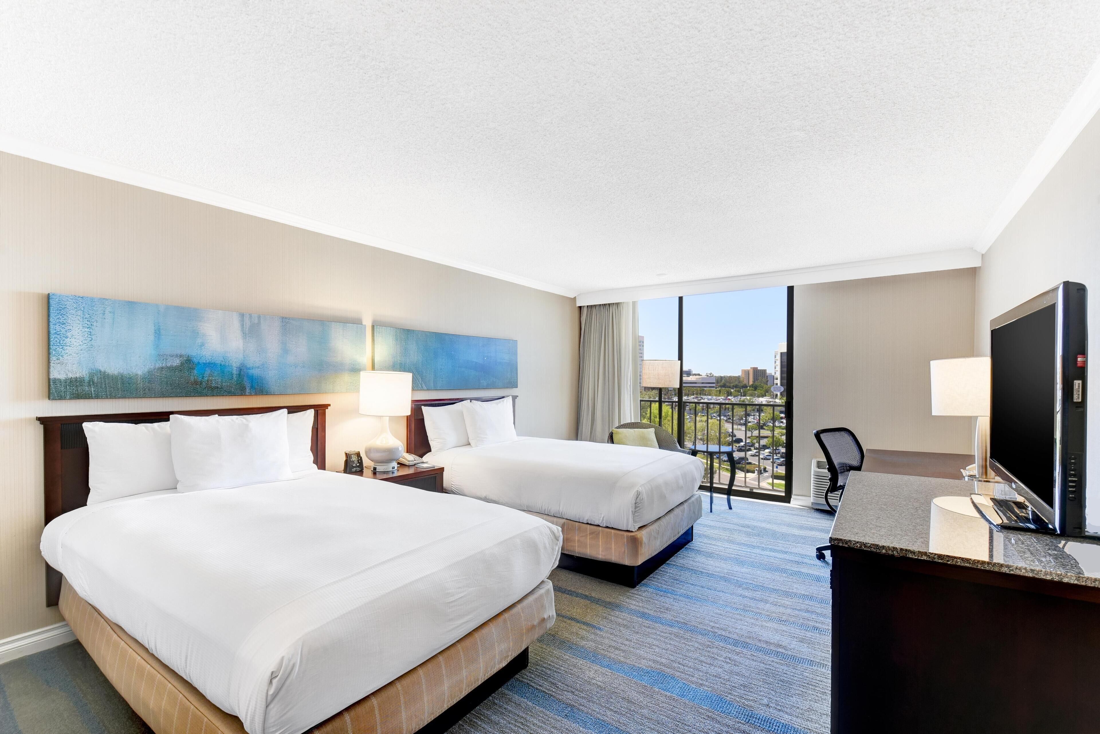 A hotel room with two beds, a desk, a television, and a large window with a view.