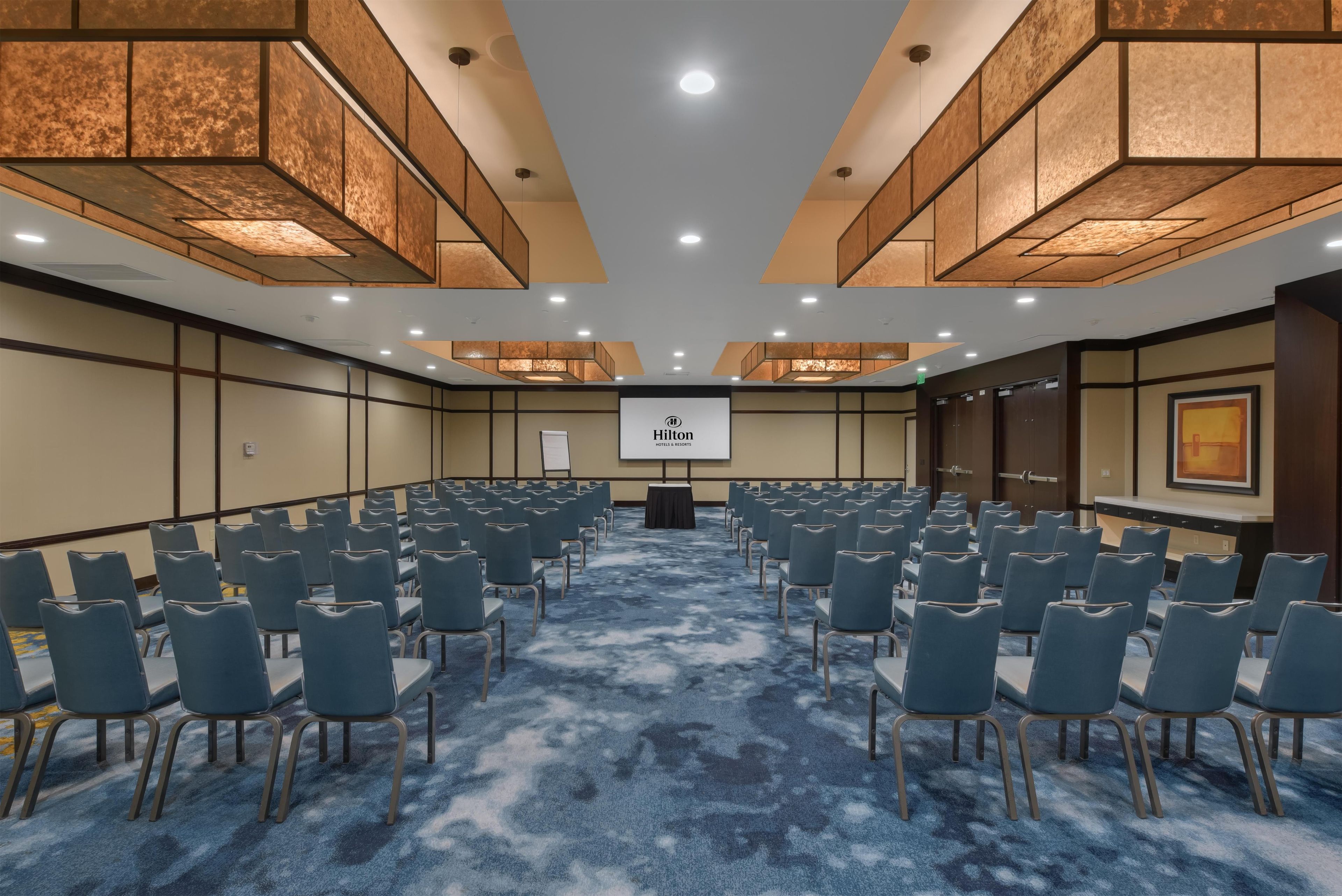 A conference room with rows of chairs, a podium, and a Hilton logo on the screen.