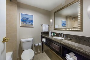 Baño con inodoro, lavamanos y espejo grande. Hay toallas colgadas en la pared y un cuadro sobre el inodoro.