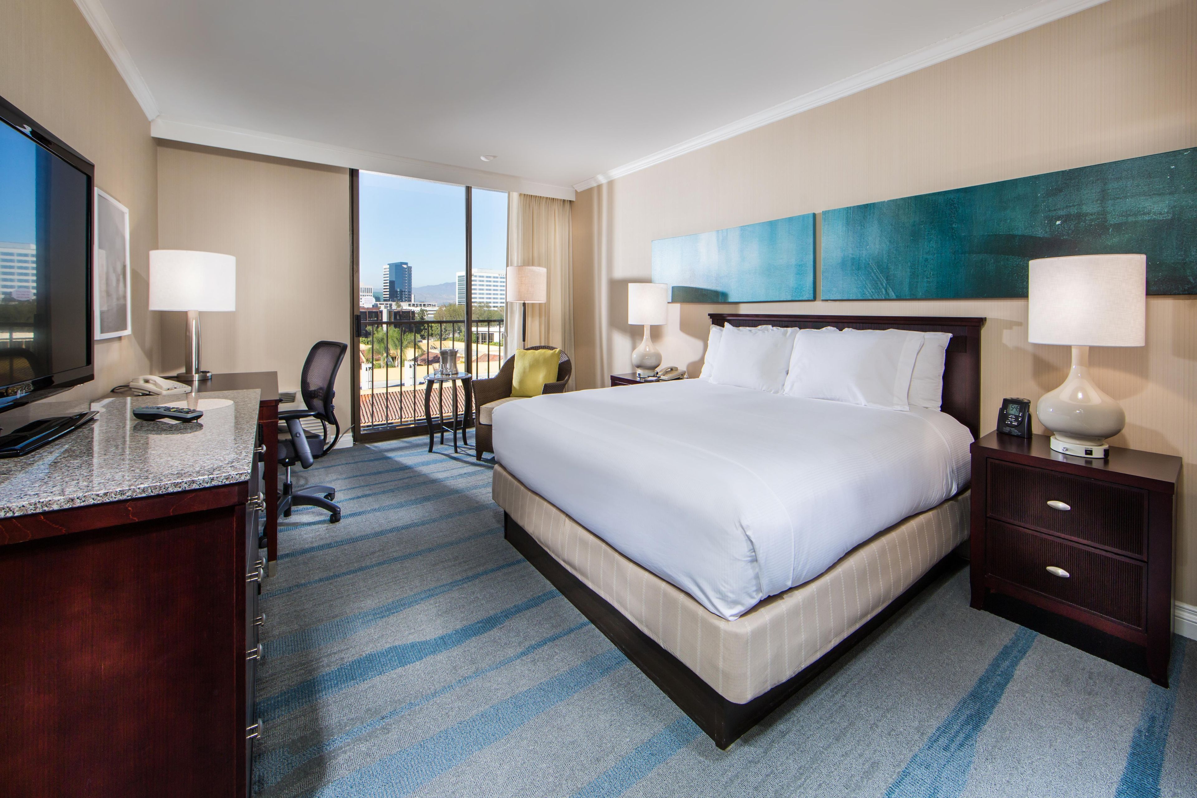 A hotel room with a large bed, a desk with a telephone, a chair, a TV, and a view of the city.
