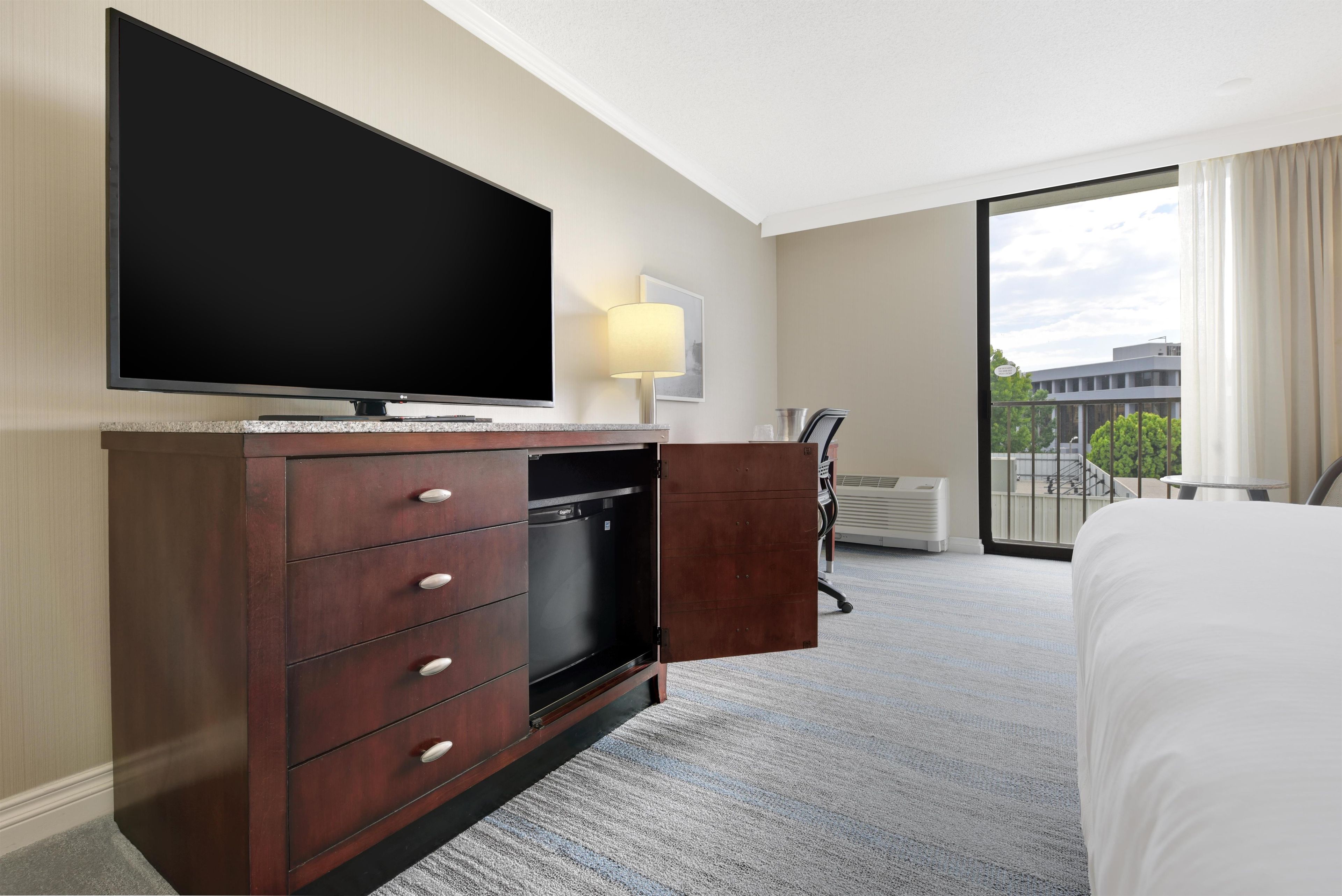 A hotel room with a large flat-screen TV, a wooden dresser with drawers, a desk with a lamp, and a large window with a view of buildings.