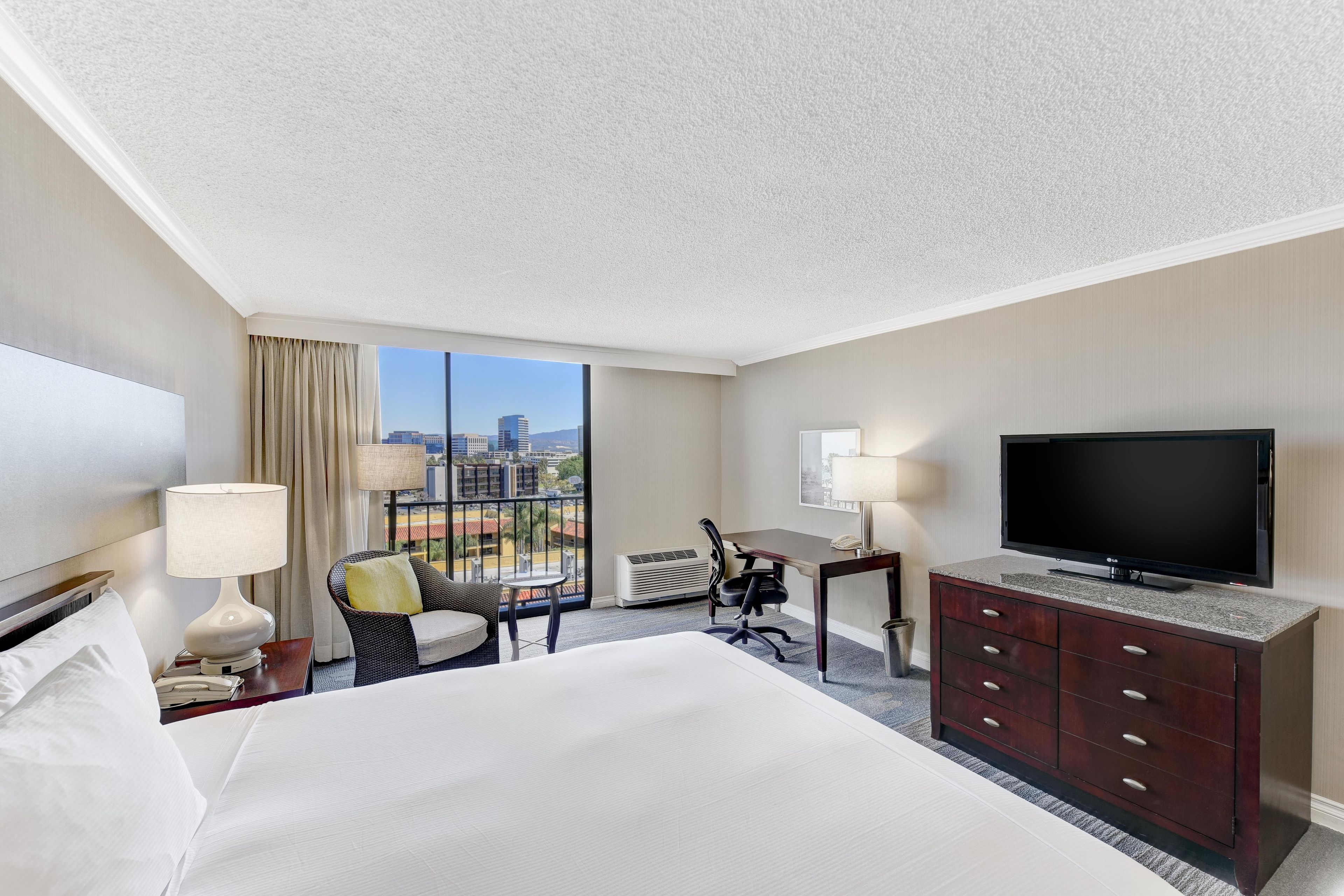 A hotel room with a bed, a desk, a television, a chair, and a view of the city.