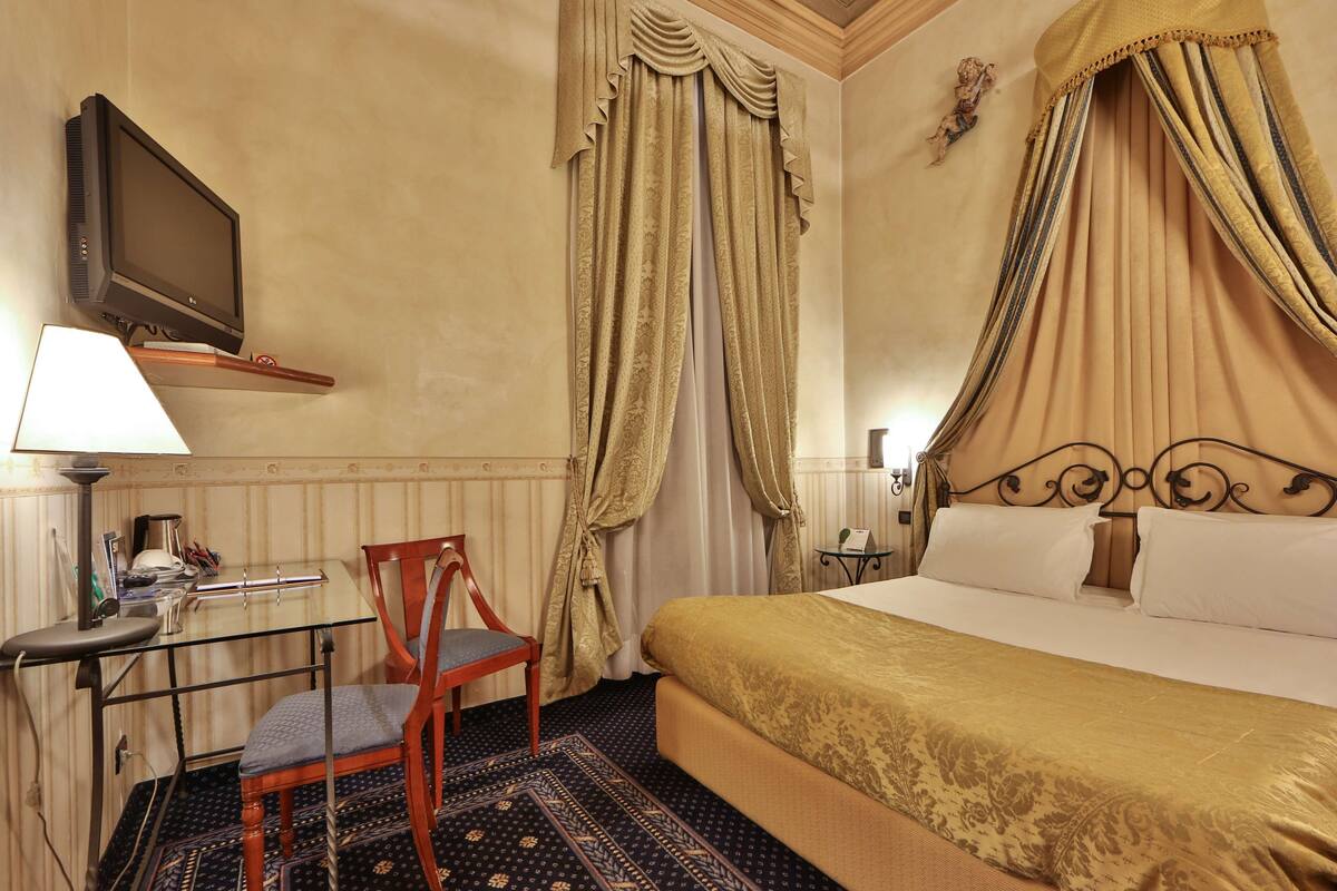 A hotel room with a bed, a desk with a chair, a TV, and a window with curtains.