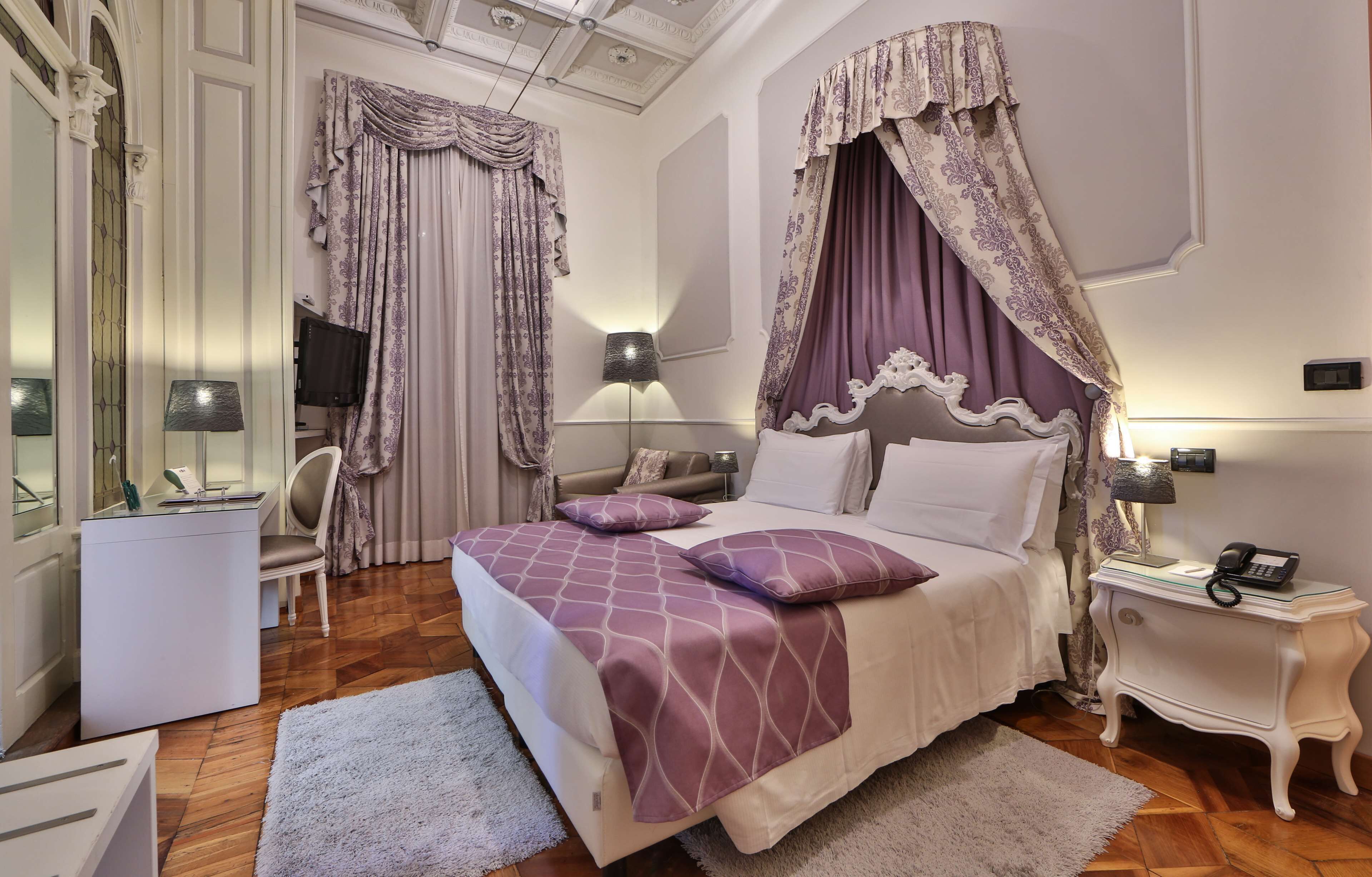 A bedroom with a canopy bed, a bedside table with a lamp, a chair, and a desk with a lamp.
