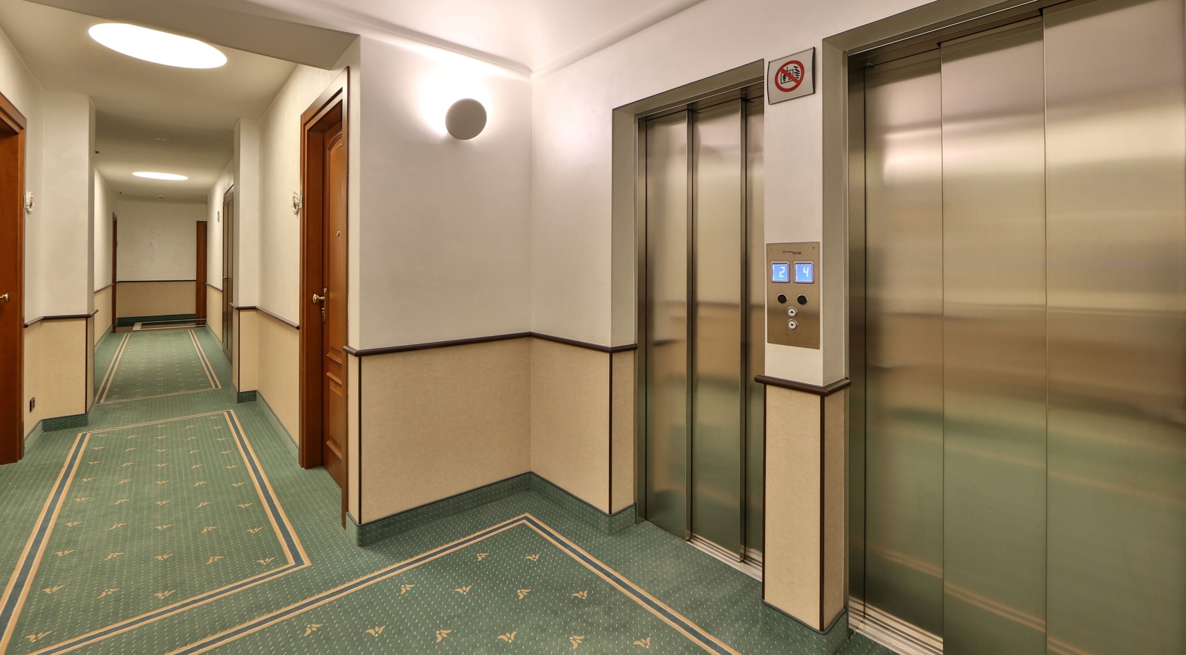 A hotel corridor with two elevators, carpeted flooring, and a no-smoking sign above one of the elevators.