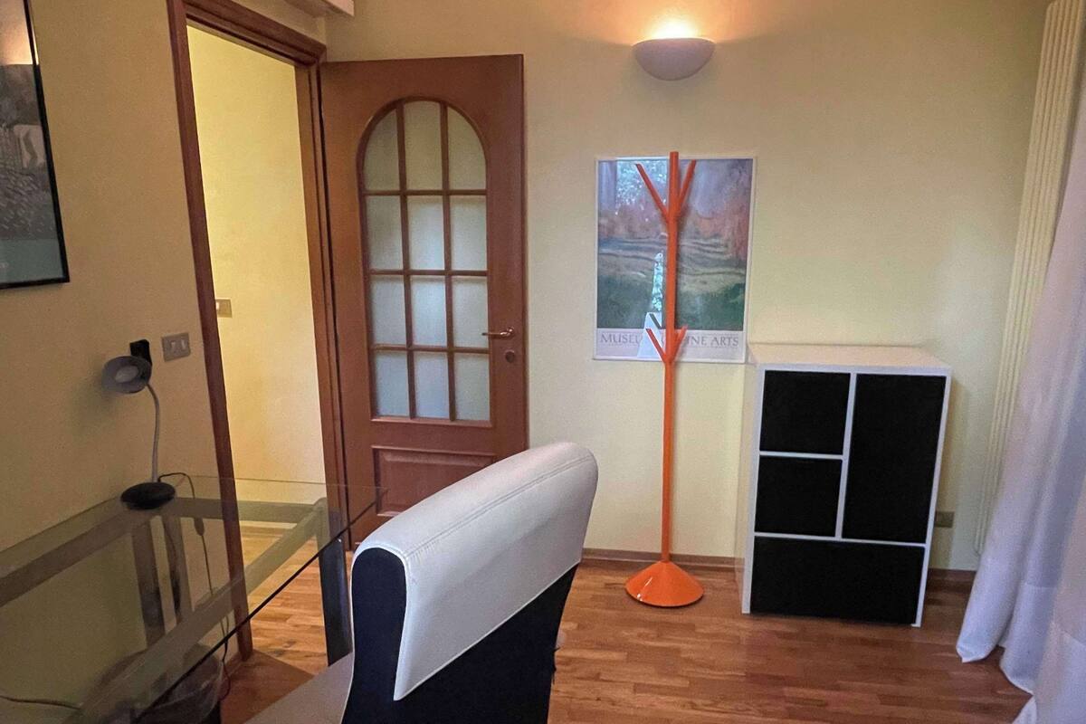 A room with a wooden floor, a desk with a chair, a door with a frosted glass panel, a wall-mounted air conditioner, a floor lamp, and a framed picture.