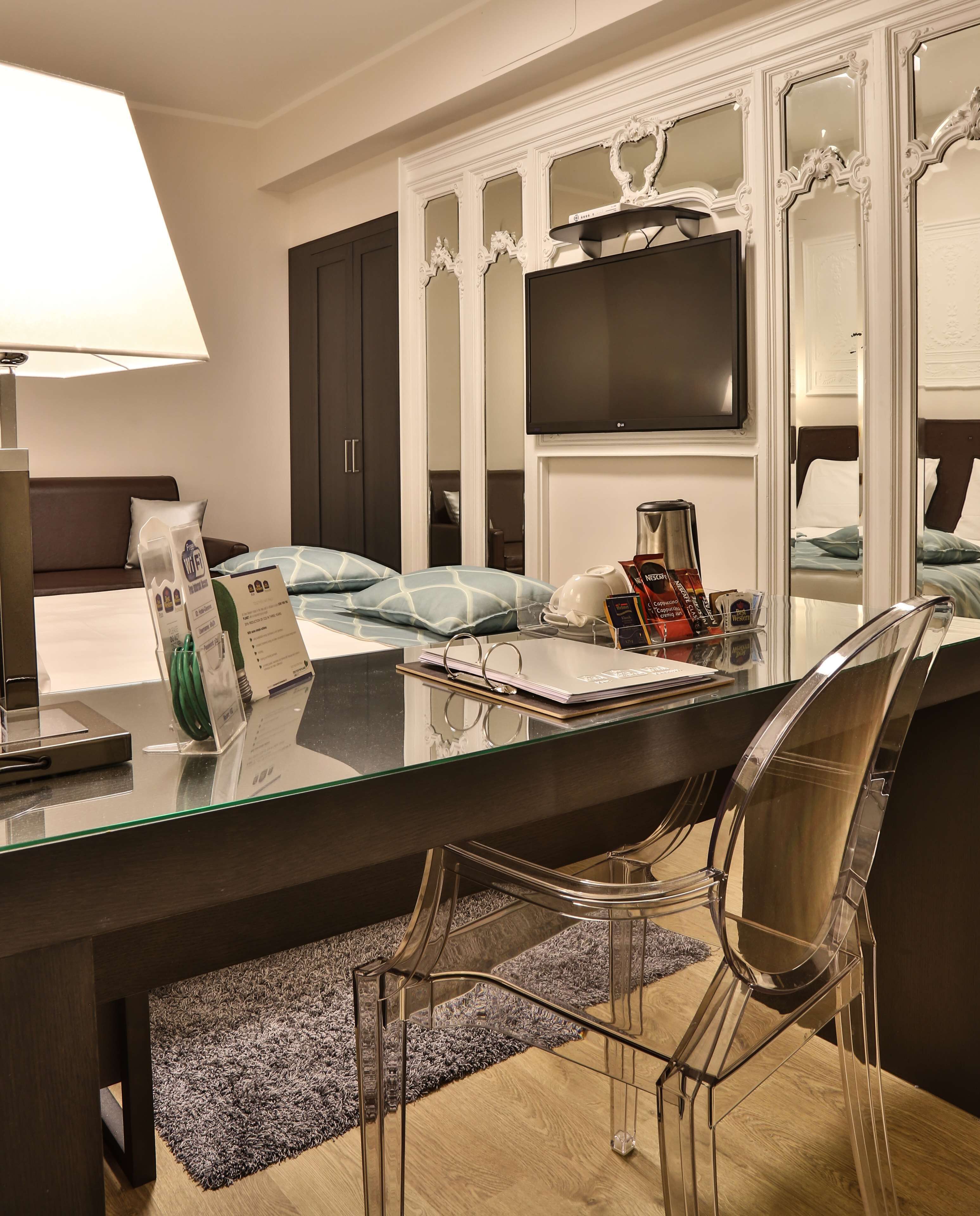 A hotel room with a glass dining table, clear chairs, a flat-screen TV, a bed with blue bedding, and a dark wooden wardrobe.