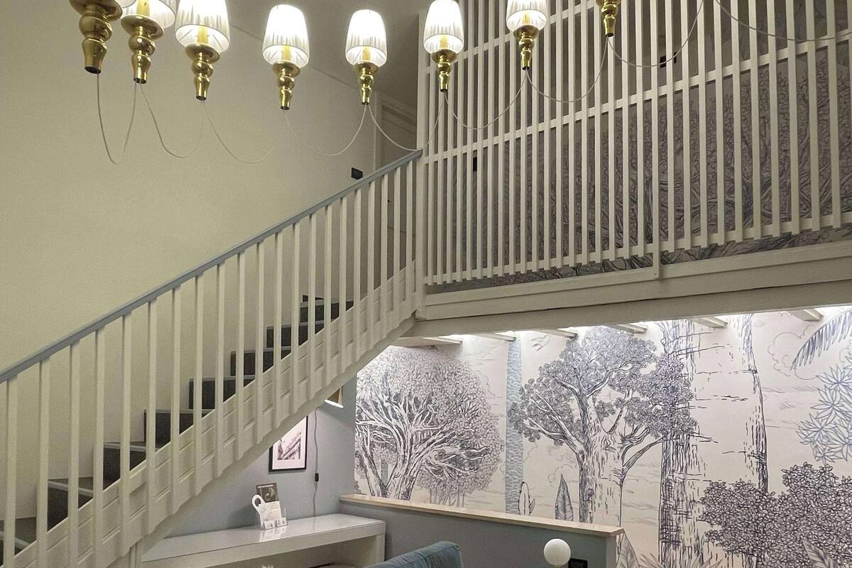 A modern living room with a grey sofa, a staircase with white railings, and a wall mural of trees.
