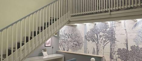 A modern living room with a grey sofa, a staircase with white railings, and a wall mural of trees.