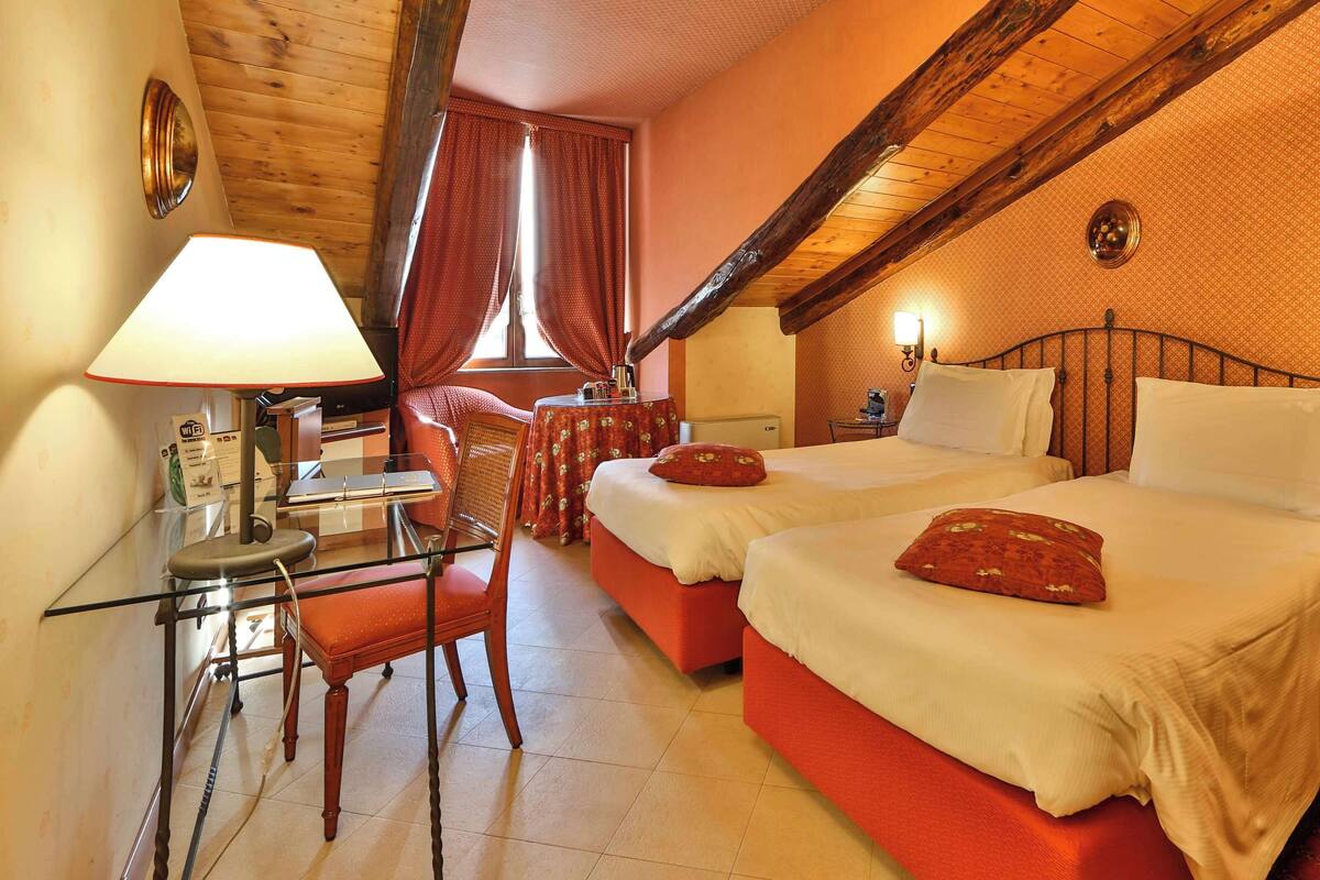 A hotel room with two beds, a desk with a lamp, a chair, and a small table.