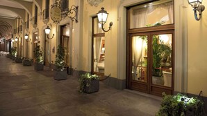 A hotel corridor with ornate wall sconces, potted plants, and a large wooden door with glass panels.