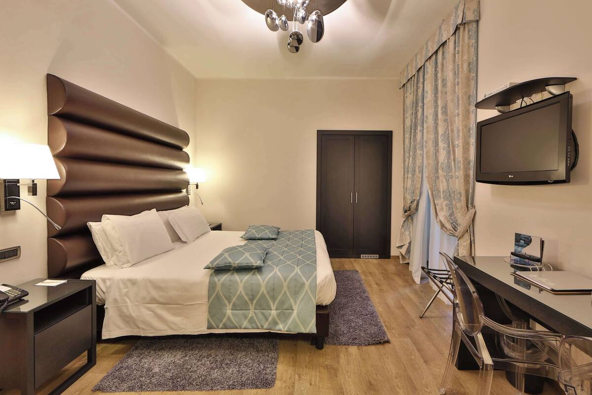 A modern hotel room with a large bed, bedside tables, a desk with a chair, a television, and a wardrobe.