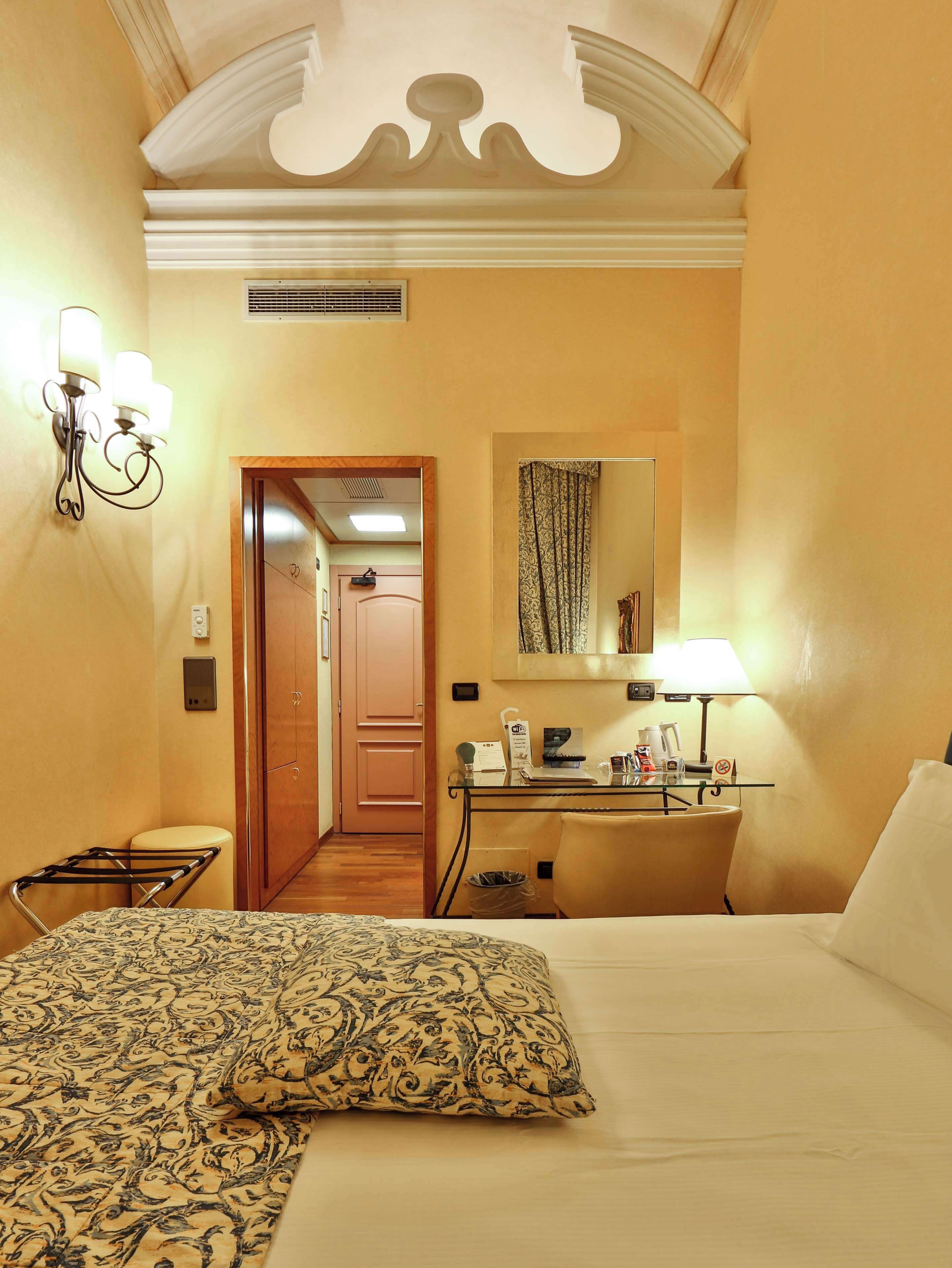 Una camera d'albergo con un letto, una scrivania con una lampada, uno specchio e una porta che dà su un'altra stanza.