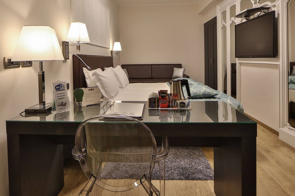 A hotel room with a bed, bedside lamps, a desk with a laptop, a chair, and a TV.