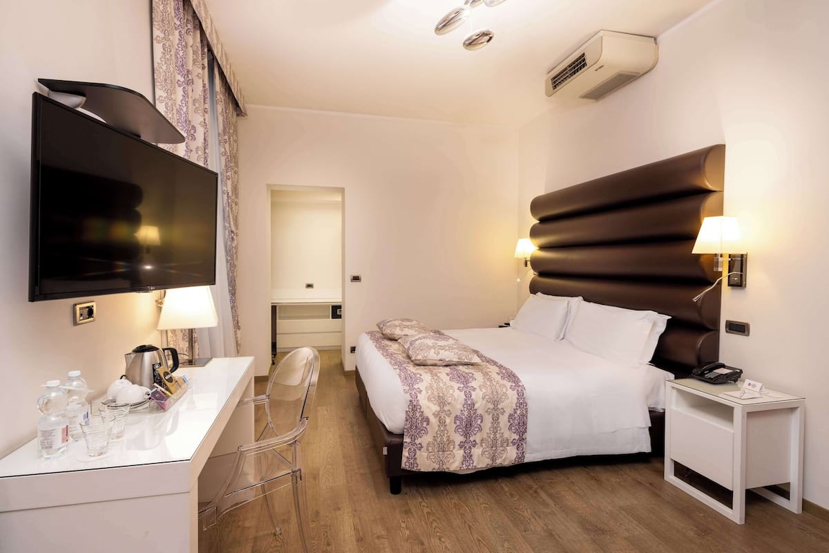 A hotel room with a bed, bedside table, lamp, TV, and a desk with a chair.