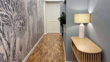 Un corridoio con pavimento in legno, un muro decorato con un murale di un albero, una porta bianca, una lampada su una consolle in legno e una pianta sullo sfondo.