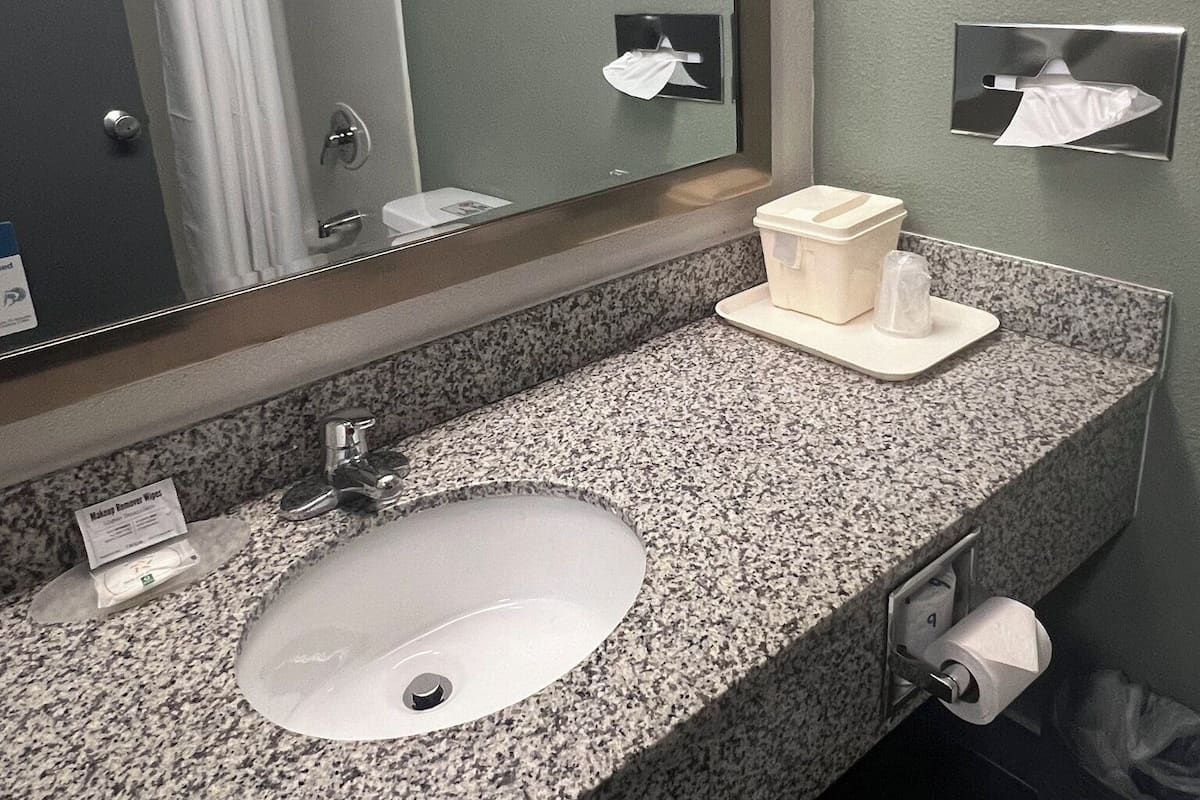 A bathroom with a granite countertop, a sink, a mirror, a towel dispenser, and a trash bin.