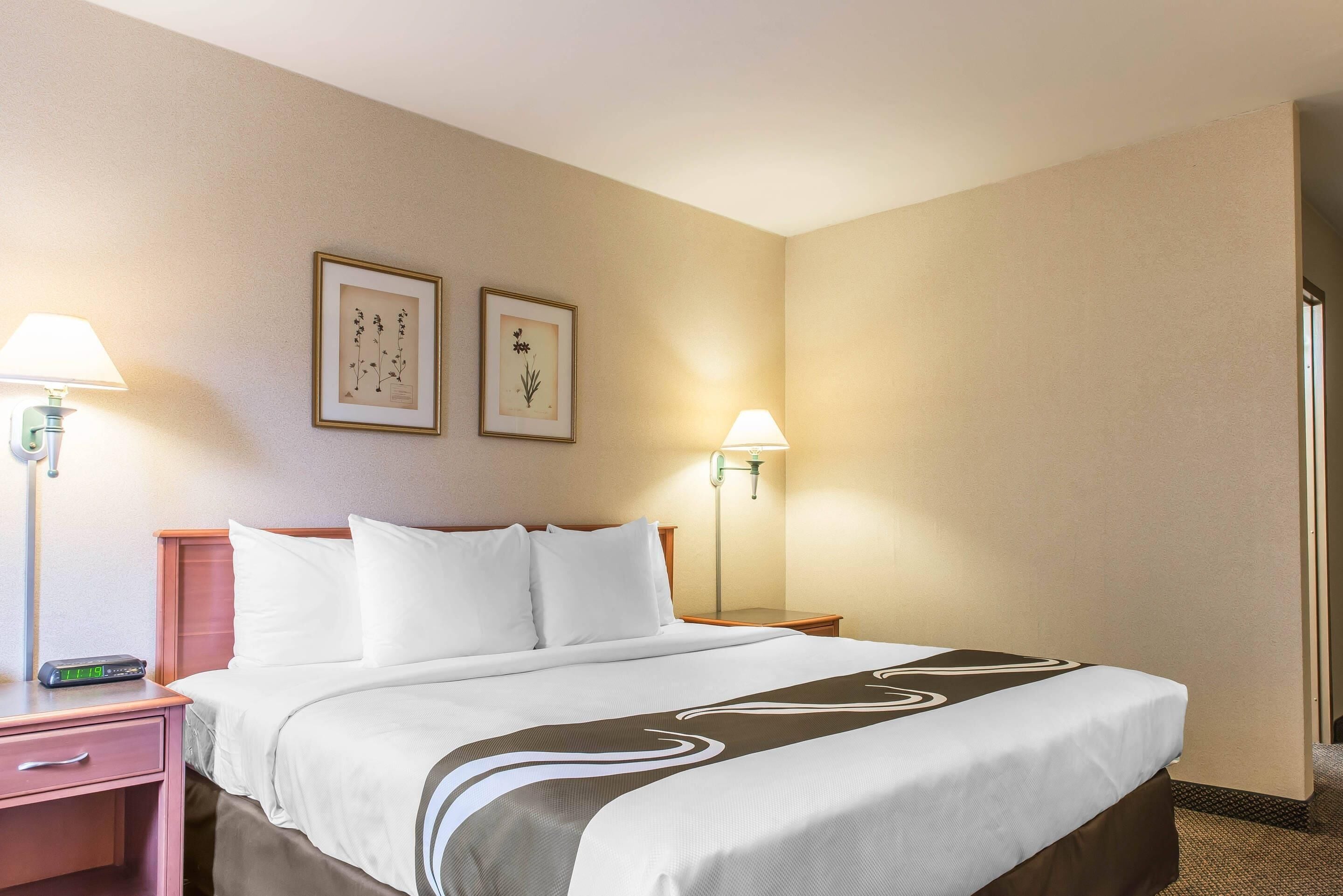 A hotel room with a bed, two bedside lamps, a nightstand, a framed picture, and a wall-mounted lamp.