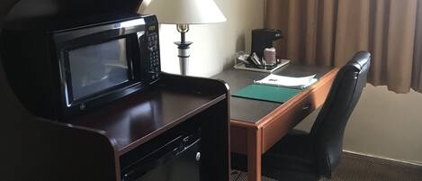 A compact hotel room with a kitchenette, a desk, a chair, a lamp, and a window with curtains.