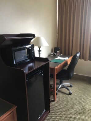 A compact hotel room with a kitchenette, a desk, a chair, a lamp, and a window with curtains.