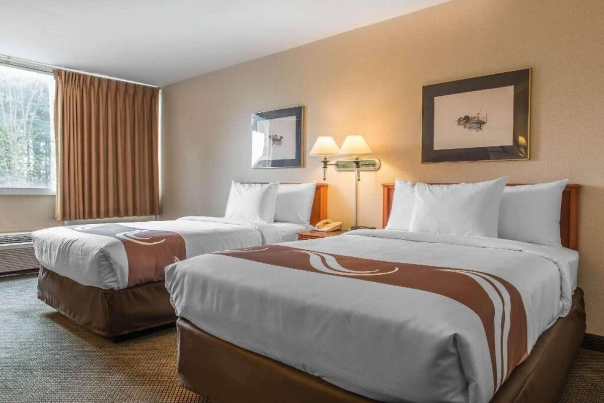 A hotel room with two beds, a window with curtains, a wall-mounted lamp, and framed artwork.