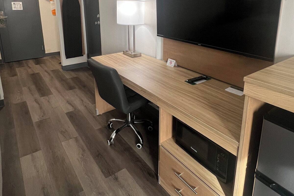 A hotel room with a wooden desk, a flat-screen TV, a lamp, a microwave, and a refrigerator.