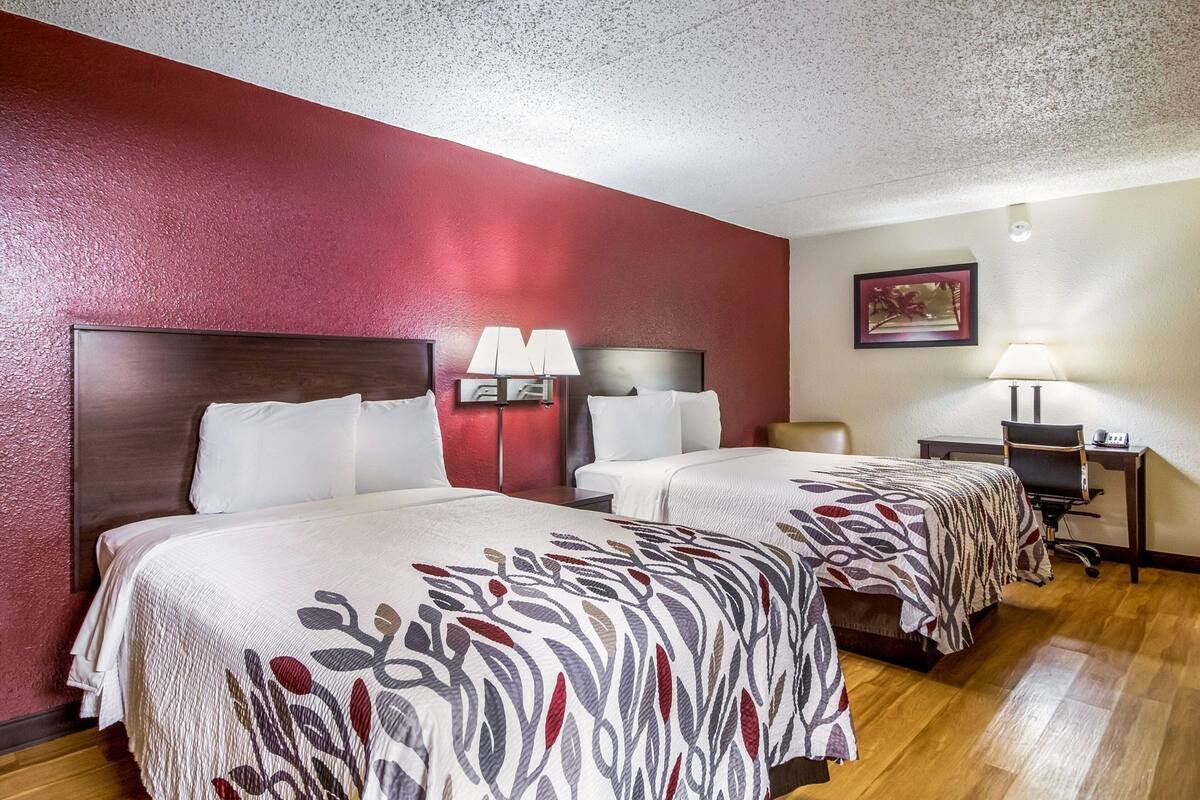 A hotel room with two beds, a desk, a chair, and a lamp.