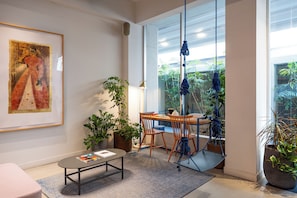 A modern living room with a large window, a painting on the wall, and a dining area with a table and chairs.