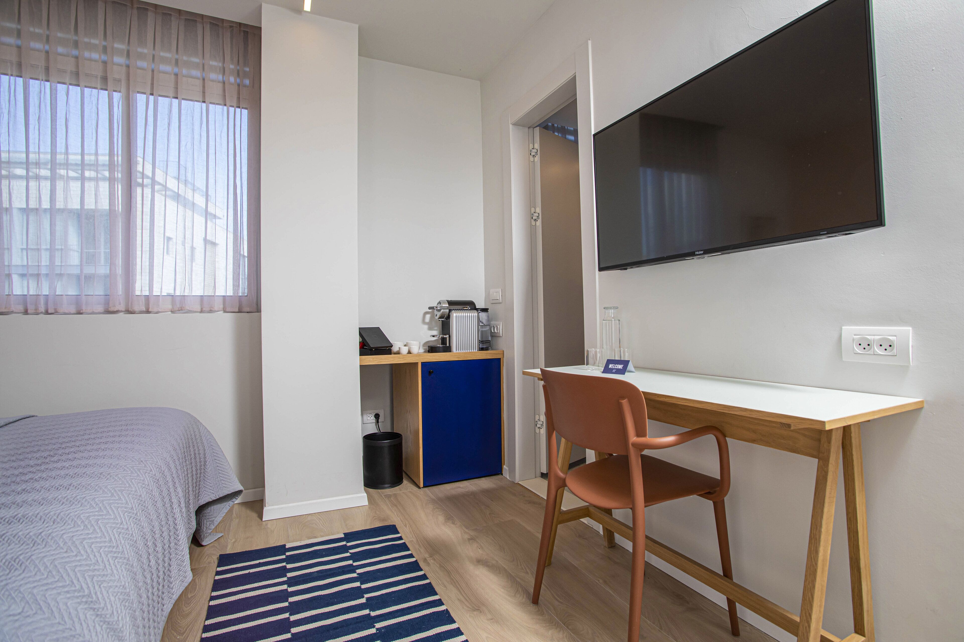 Habitación compacta con cama, escritorio, silla, kitchenette pequeño y televisor montado en la pared.