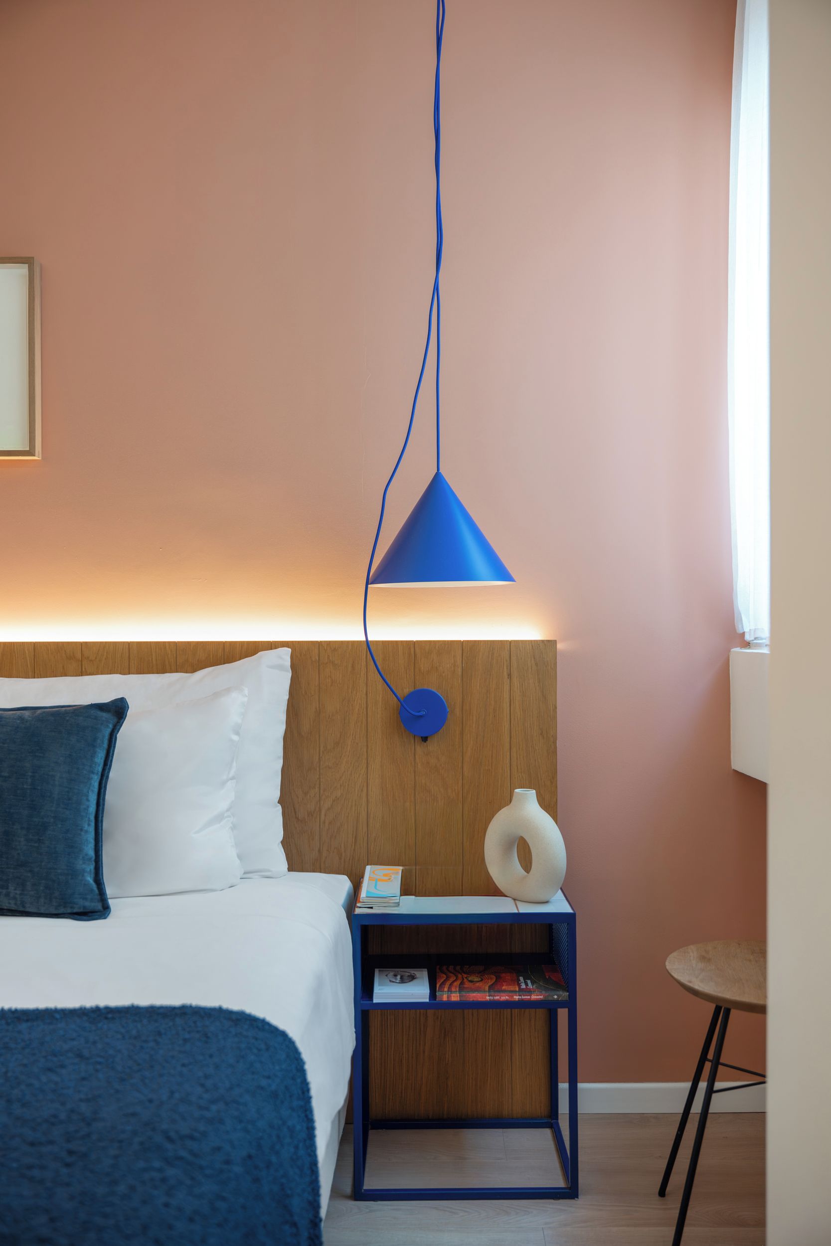 A hotel room with a bed, a bedside table with a lamp, a small round table, and a wooden headboard.