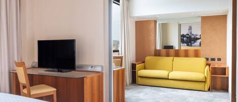 A hotel room with a bed, a desk with a TV, a yellow sofa, and a large window.