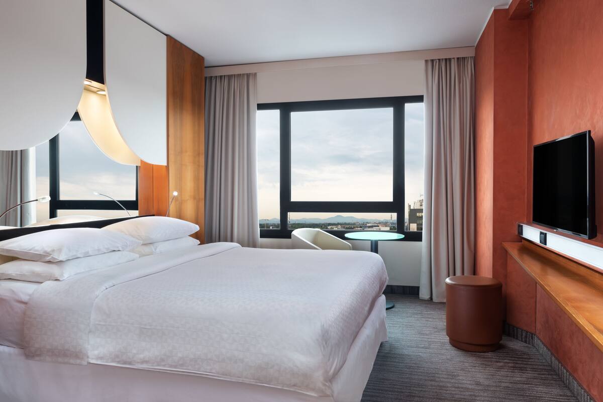 A hotel room with a large bed, a TV, a small table, and a view of the city through large windows.