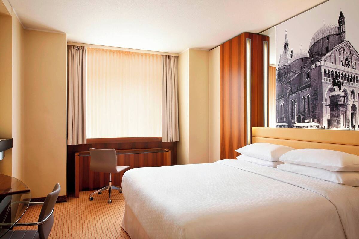 A hotel room with a large bed, a desk with a chair, a window with curtains, and a wall-mounted artwork.