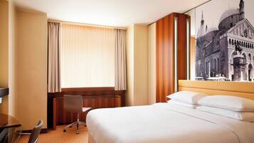 Habitación de hotel con una cama grande, un escritorio con silla, una ventana con cortinas y una obra de arte colgada en la pared.