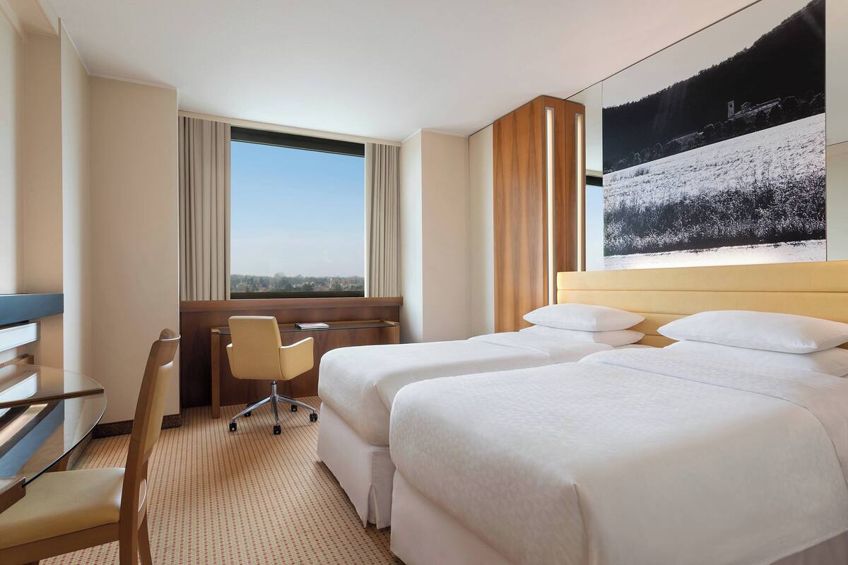 A hotel room with two beds, a desk, a chair, and a large window offering a view of the city.