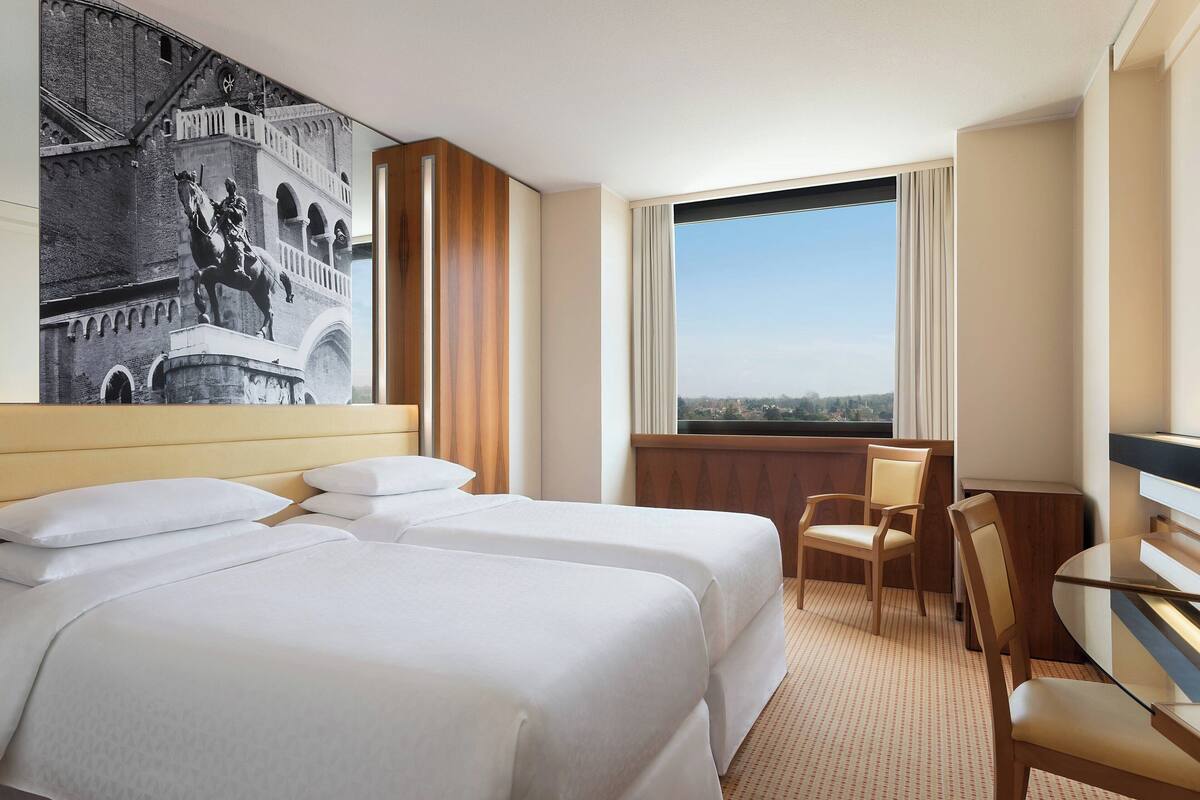 A hotel room with two beds, a desk, and a large window offering a city view.