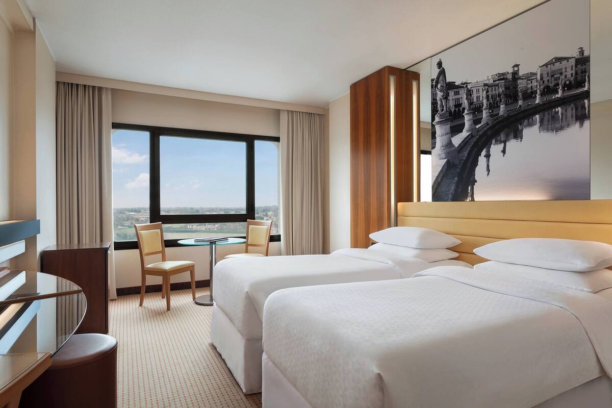 A hotel room with two beds, a desk, a chair, and a large window offering a city view.