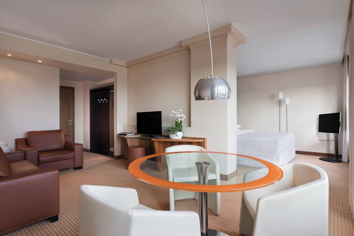 A modern hotel room with a glass table, leather sofas, a flat-screen TV, a bed, and a large window.
