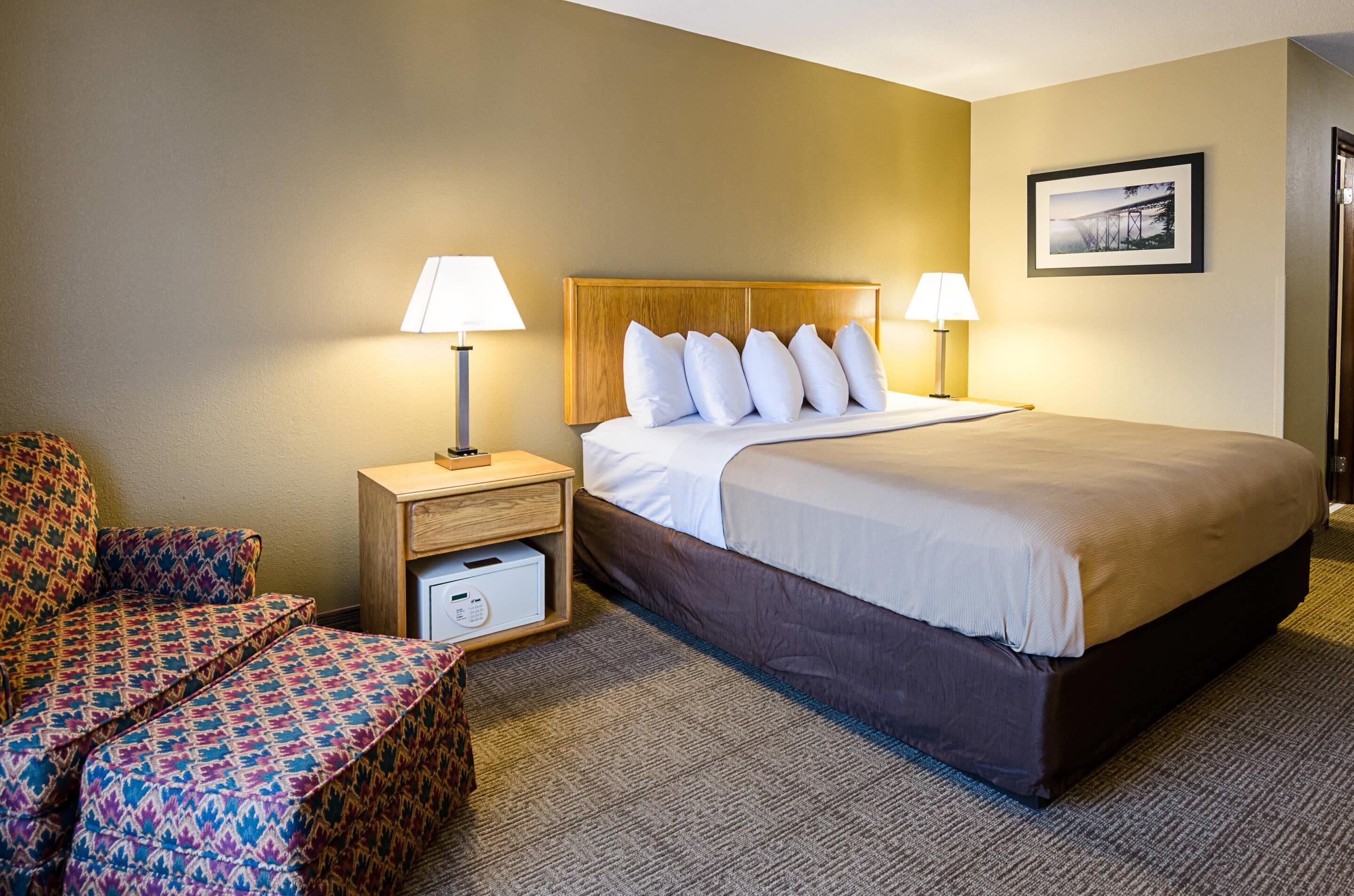 A hotel room with a large bed, two bedside lamps, a wooden nightstand, a patterned armchair, and a framed picture on the wall.