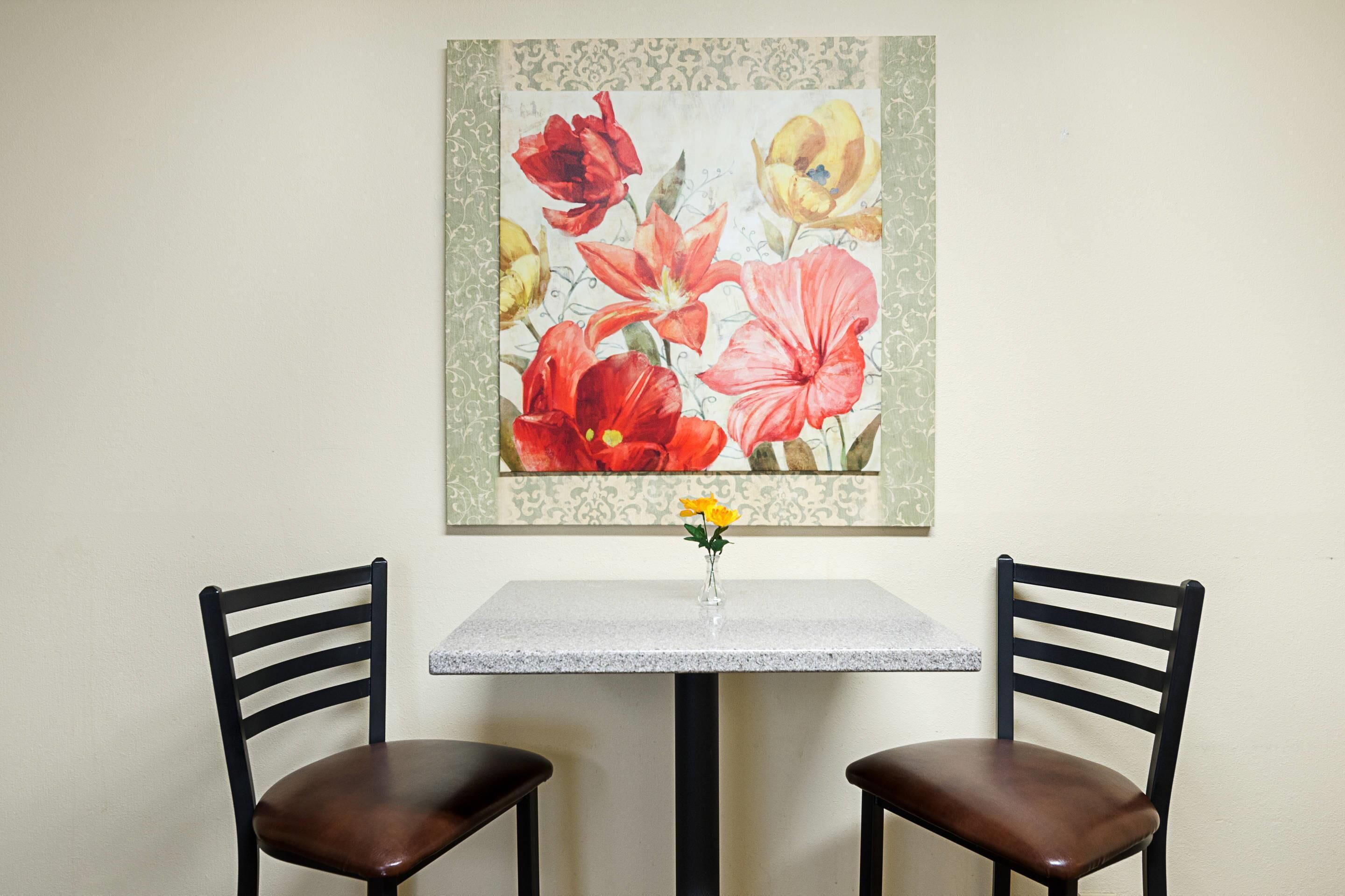 Un comedor con una mesa y dos sillas, un cuadro floral en la pared y un jarrón con flores amarillas sobre la mesa.