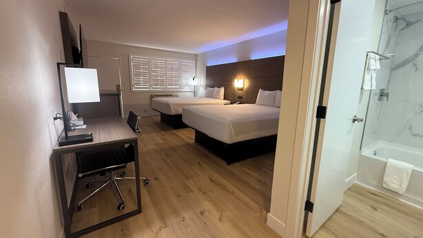 A hotel room with two beds, a desk with a computer, a desk lamp, a chair, a window with blinds, and a bathroom with a shower and bathtub. - Travelodge by Wyndham Imperial/El Centro (Imperial)