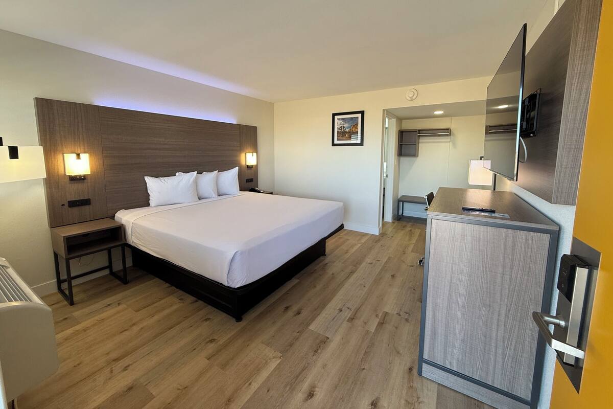 A hotel room with a large bed, bedside tables, a flat-screen TV, and a small kitchenette.