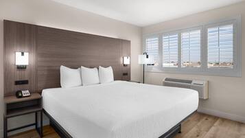 Una cama grande con ropa de cama blanca, cabecera de madera, lámparas de noche, mesita de noche con teléfono y una ventana con persianas.