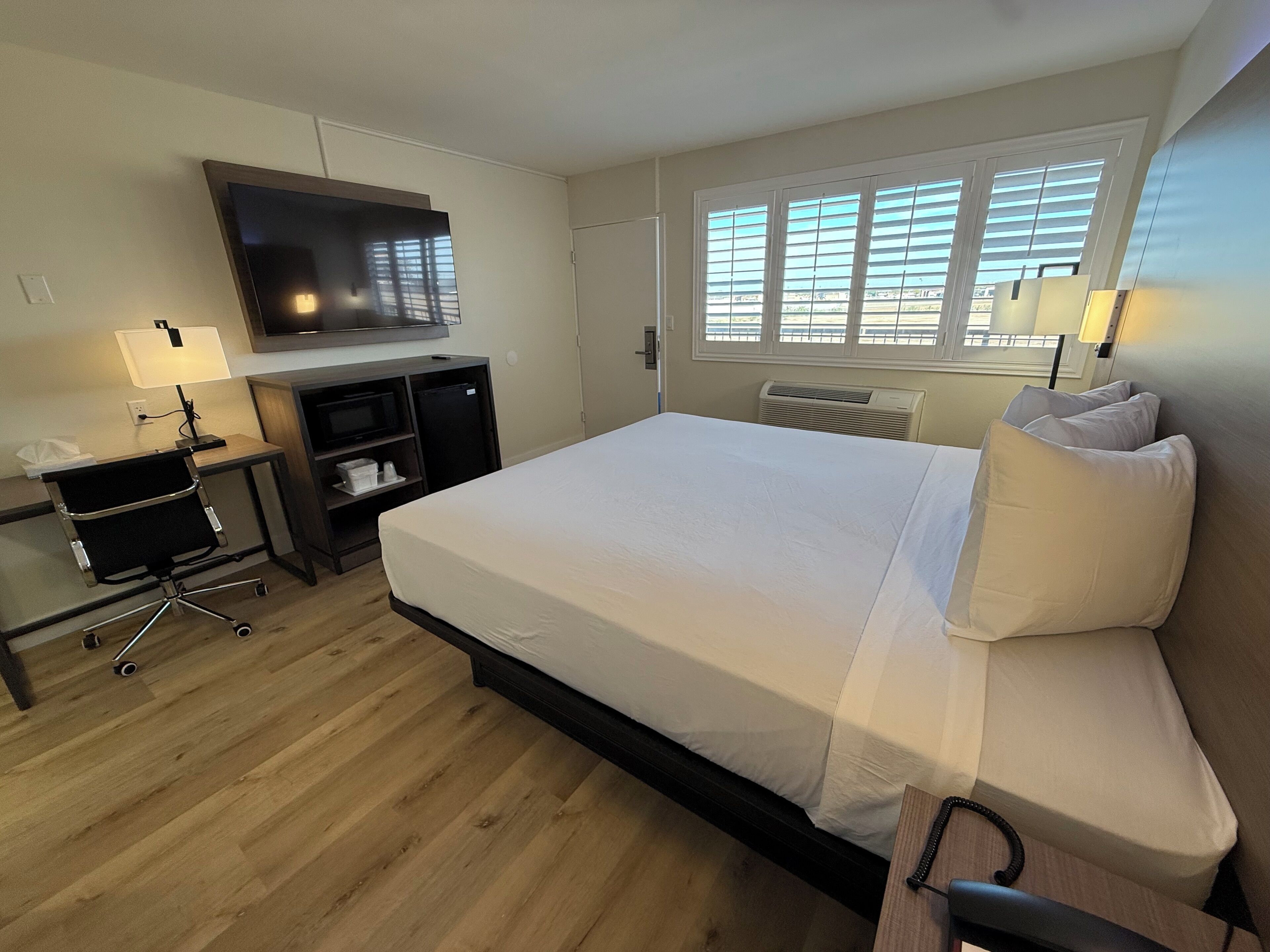 A hotel room with a bed, desk, chair, TV, and windows.