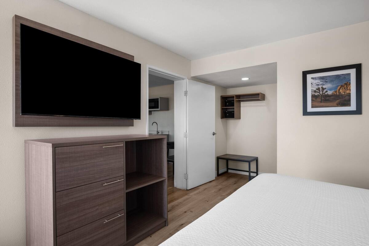 A bedroom with a bed, a TV mounted on the wall, a wardrobe, a small table, and a framed picture on the wall.