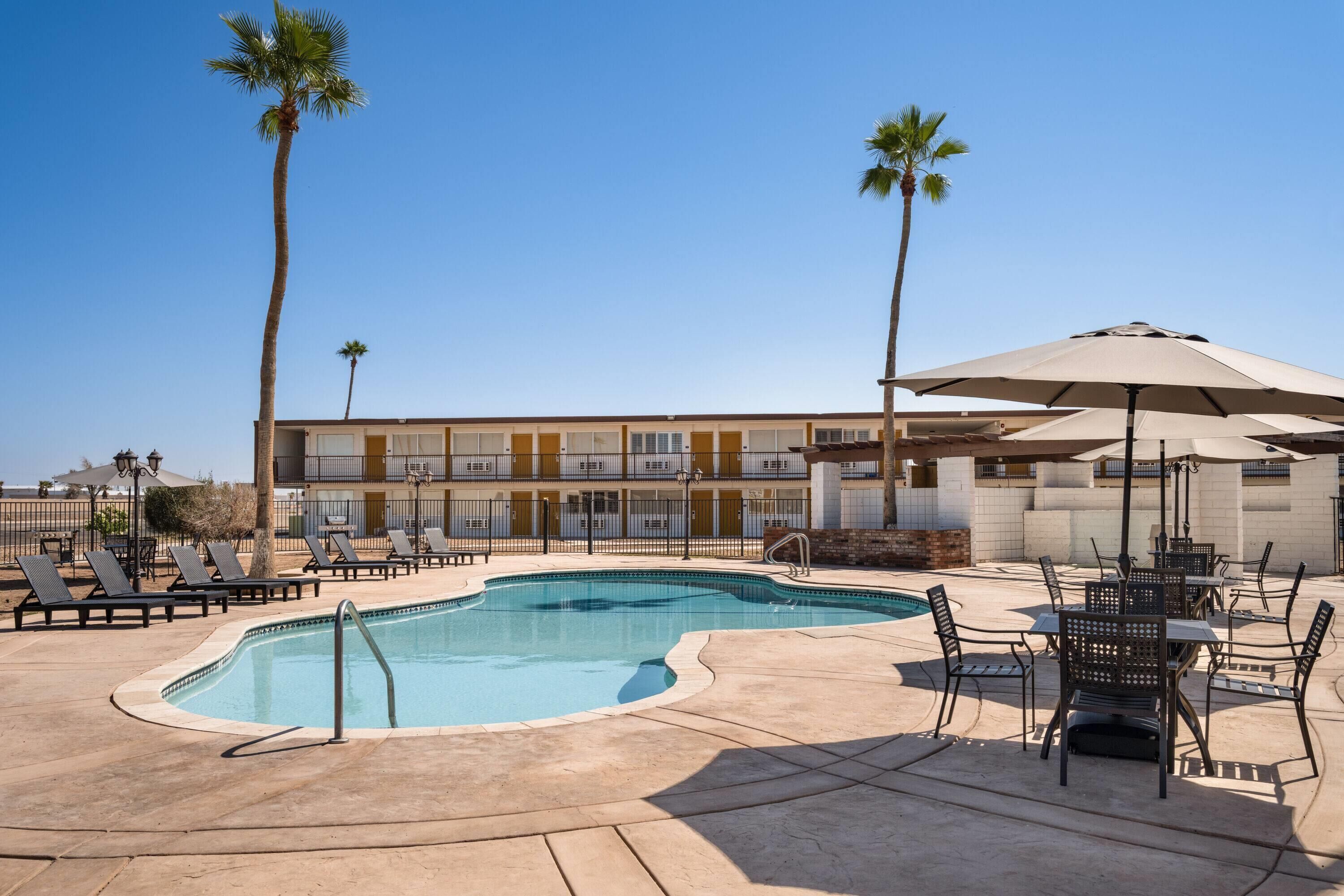 A swimming pool with lounge chairs and umbrellas, a building with balconies, and palm trees.
