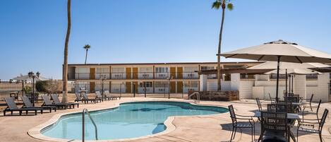A swimming pool with lounge chairs and umbrellas, a building with balconies, and palm trees.